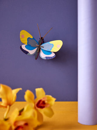 A yellow and blue cardboard monarch butterfly., mounted on a purple wall with yellow flowers in the left foreground.
