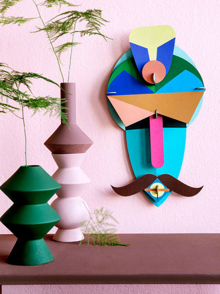 Two vases with plants on the left, and on the wall to the right of them, A colorful carboard mask depicting a man with a turban and mustache.