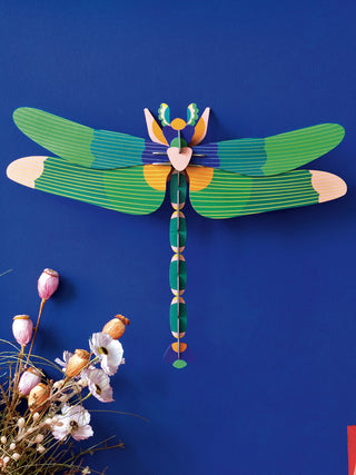 A large, cardboard green dragonfly mounted on a blue wall with  pink and orange flower bulbs in the lower left.