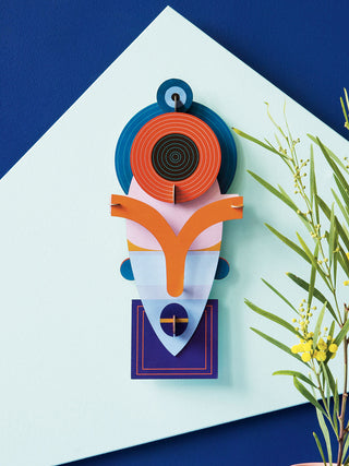 Against a light and dark blue wall, and adjacent to a plant, A colorful cardboard mask with an angular bottom and round top.