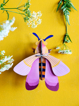 A pink and orange cardboard bee with blue antenna, mounted on a yellow wall, surrounded by plants.