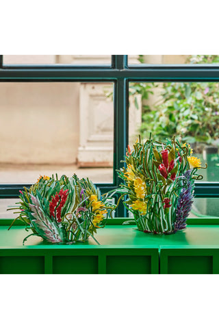 On a green shelf are two vases that look like they are made by nature, with red, yellow and purple flowers popping up among what appear to be green grasses and plants.