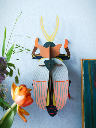 Mounted on a blue wall, adjacent to flowers, A large cardboard beetle with a blue and red body, orange head covering and yellow antenna.