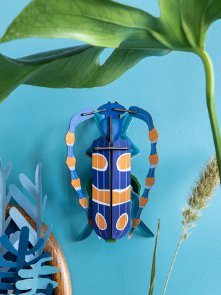 A blue and yellow cardboard beetle mounted on a light blue wall, surrounded by plants,  with antenna that curve around its body.
